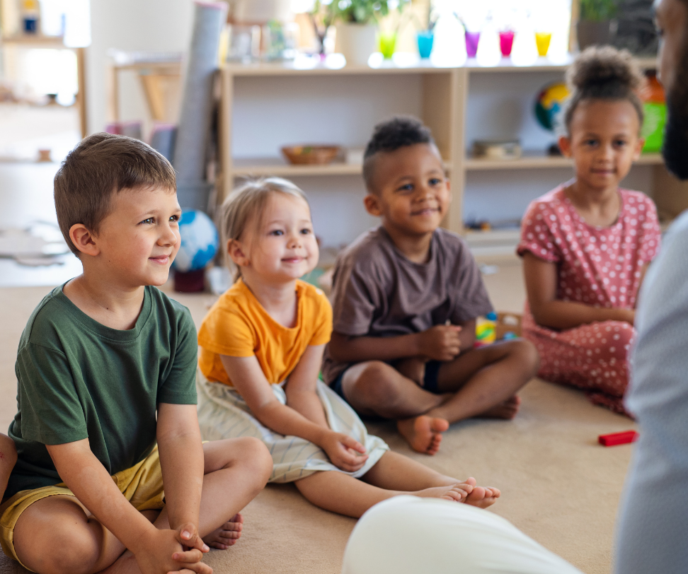 A group of children are sitting on the floor in a circle listening to a teacher.