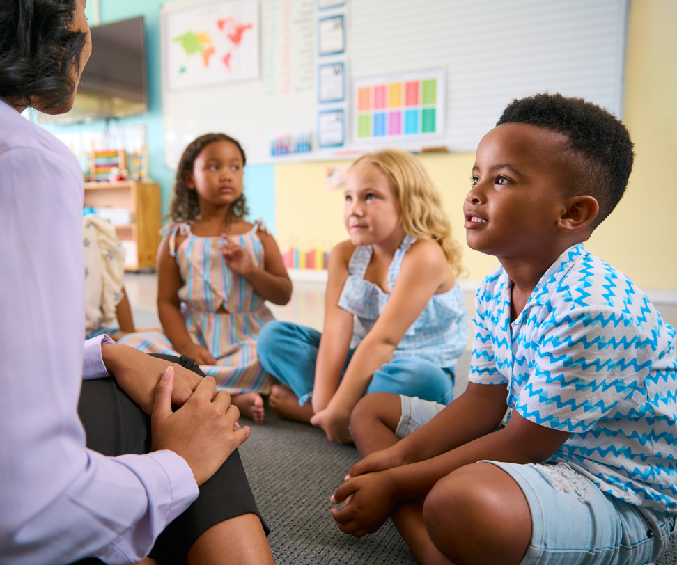 A group of children are sitting on the floor in a circle listening to a teacher.