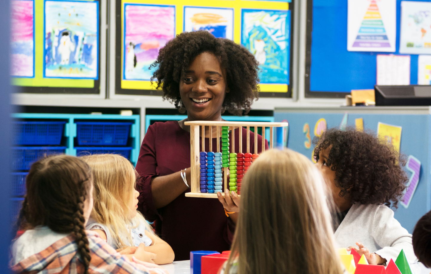 A teacher is holding a wooden abacus in front of a group of children in a classroom.