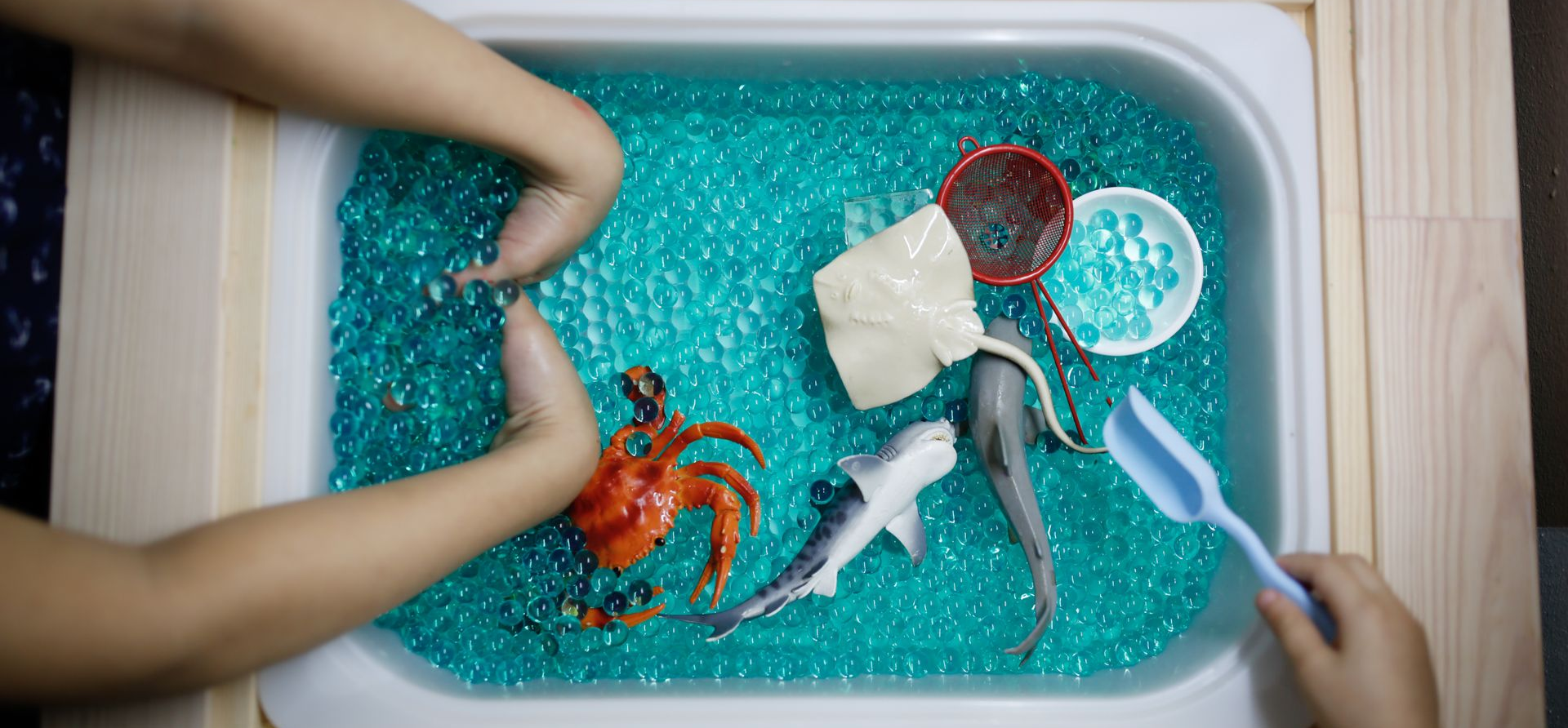 Child's hands playing with toy sea creatures in a tray of blue gel beads.