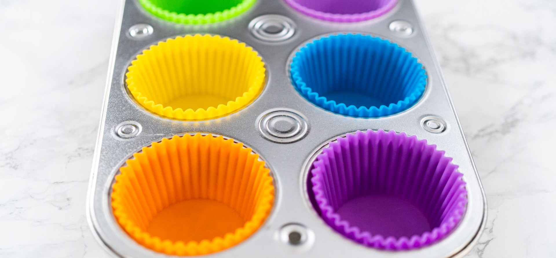 Muffin tin with colorful silicone liners. Orange, yellow, blue, green and purple cups sit in a metallic pan.