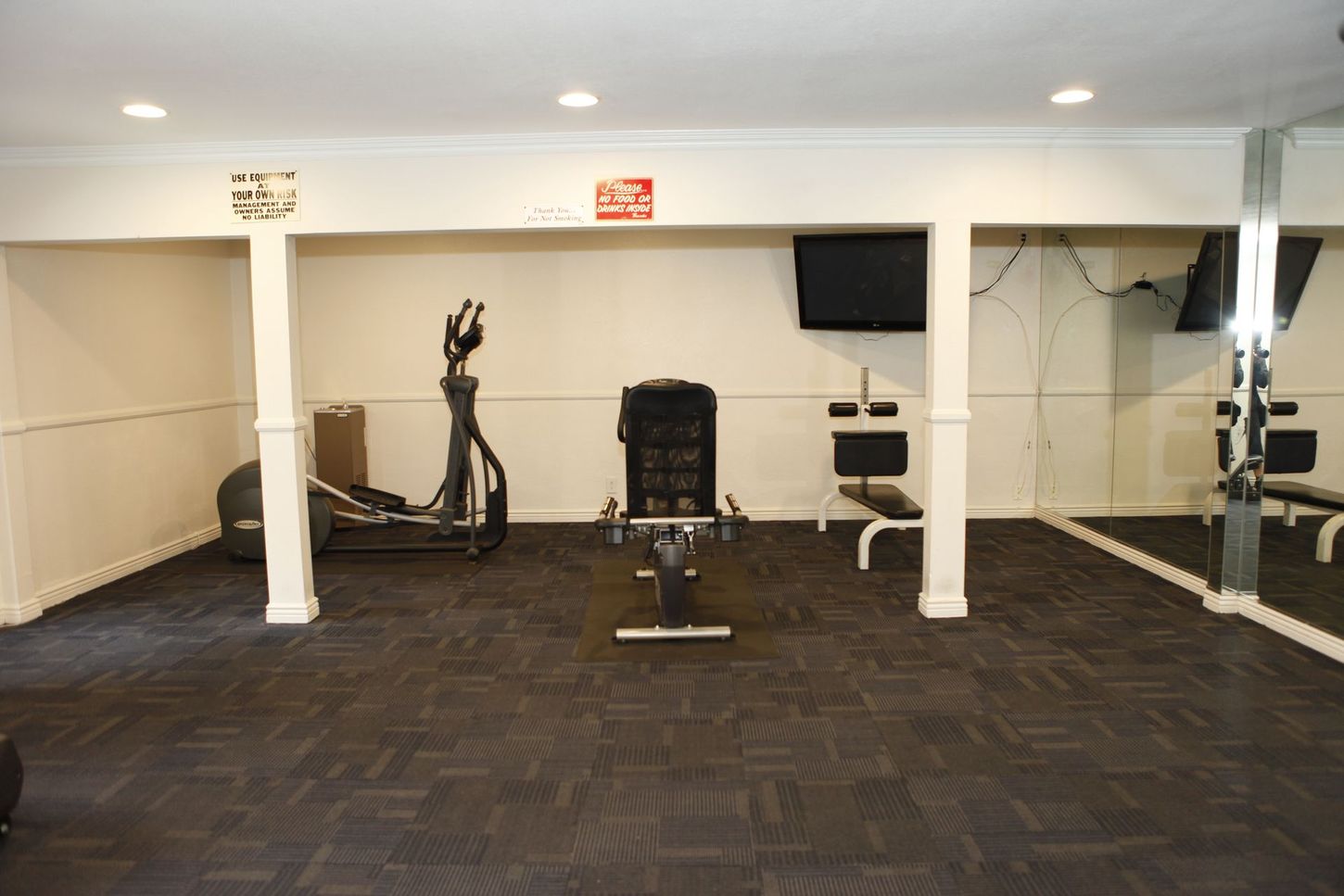 Home gym with elliptical machine, weight bench, and TV. Black rubber flooring, white walls.