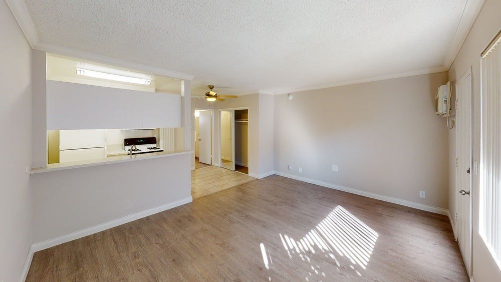 Empty apartment interior with kitchen, wooden floors, and sunlight through blinds.