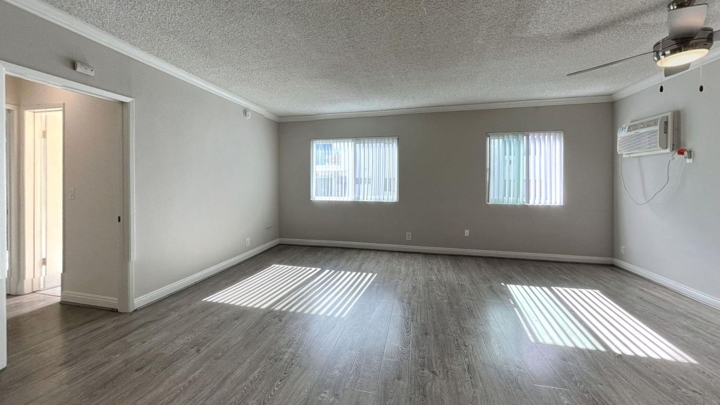 Empty living room with gray walls, wood-look floor, windows, and ceiling fan.