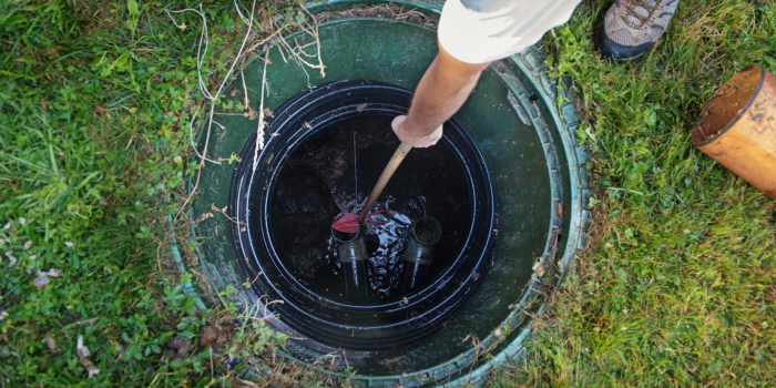 Person using a tool to stir contents of a septic tank. Surrounded by green grass.