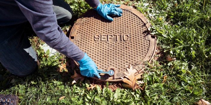 Person opening a septic tank cover in a grassy area, wearing blue gloves.