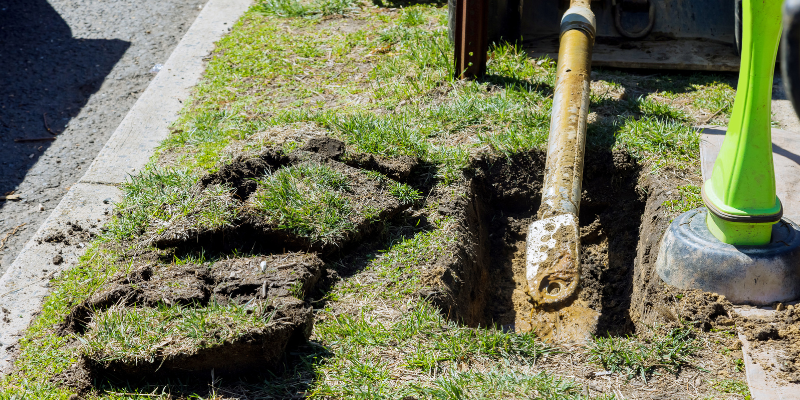 A shovel in a hole dug in a grassy area, with cut sod pieces nearby, next to a paved area.