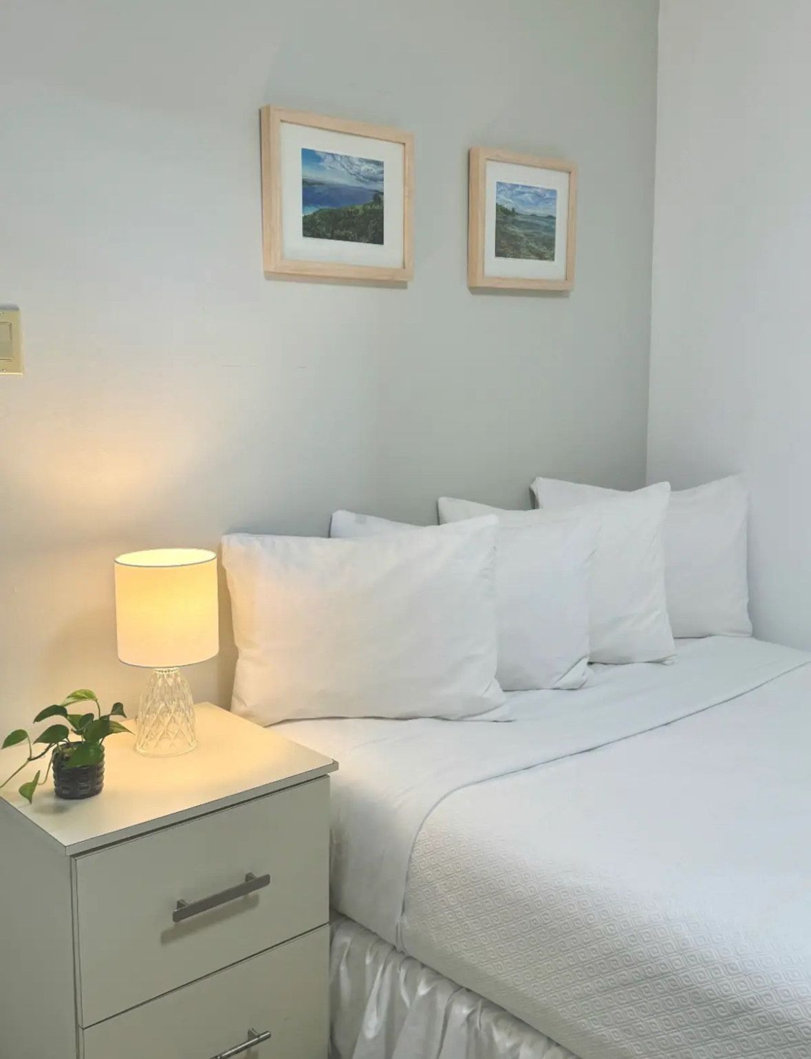 A bedroom with a bed , nightstand , lamp and two pictures on the wall.