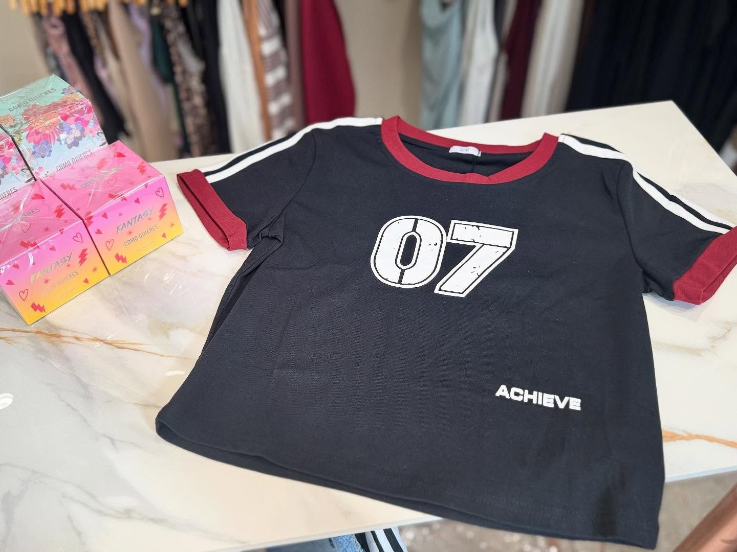 Camiseta negra con texto "07" y "ACHIEVE" en blanco, ribete rojo y rayas blancas en las mangas, sobre superficie de mármol.
