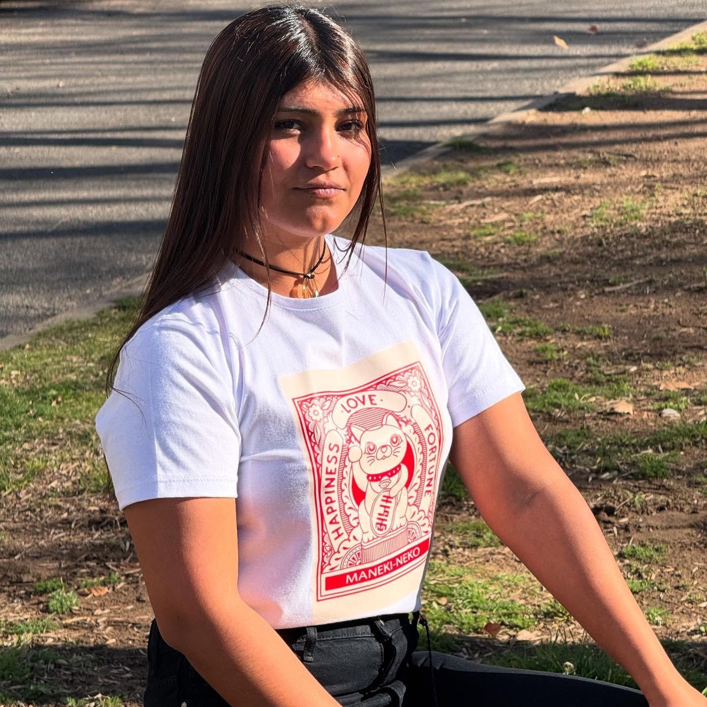 Mujer con cabello largo y oscuro, con un top blanco con gráficos rojos, sentada afuera.