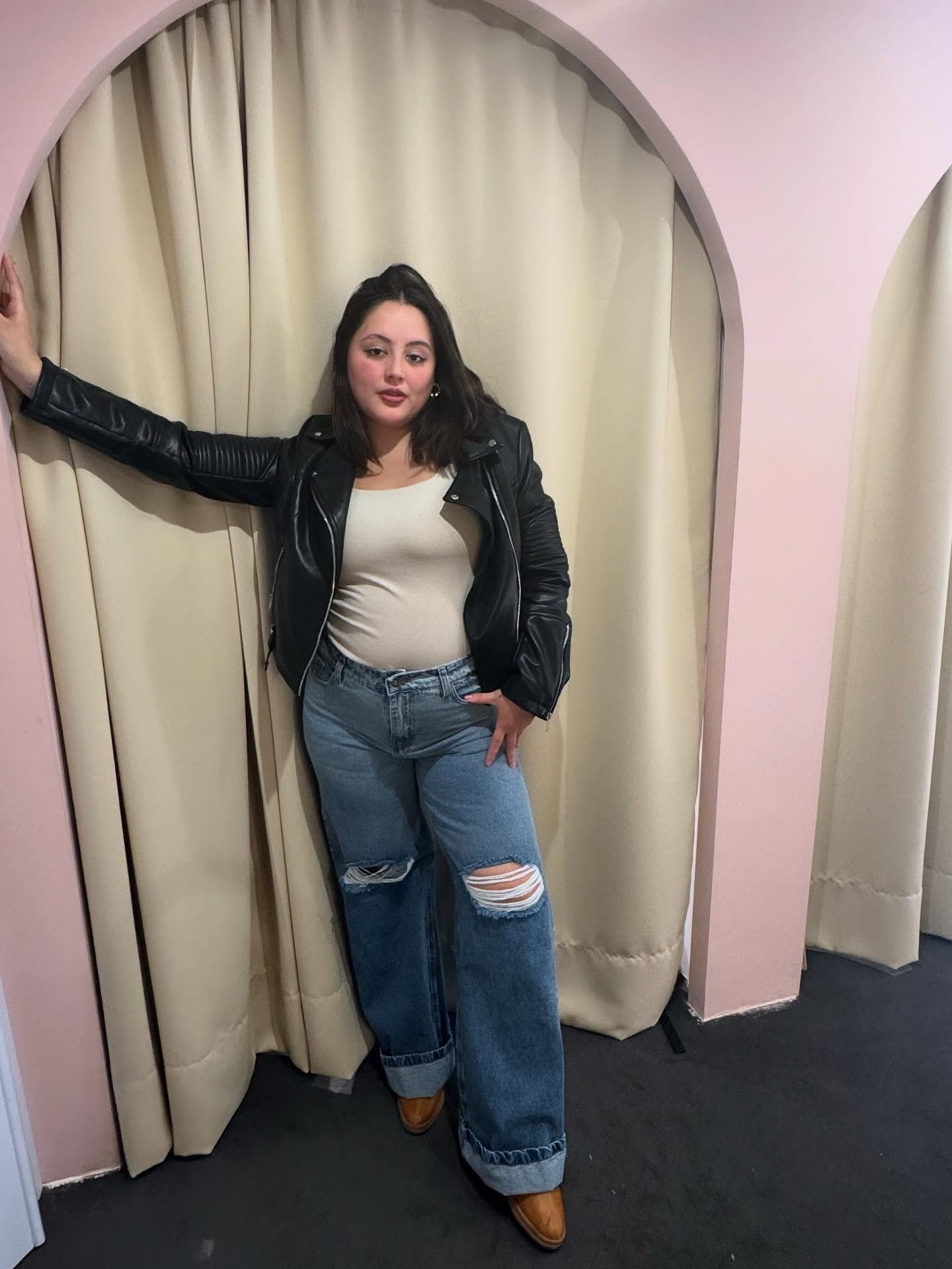 Una mujer con una chaqueta negra y pantalones vaqueros rotos posa junto a una pared con cortinas; arcos rosas en una tienda.