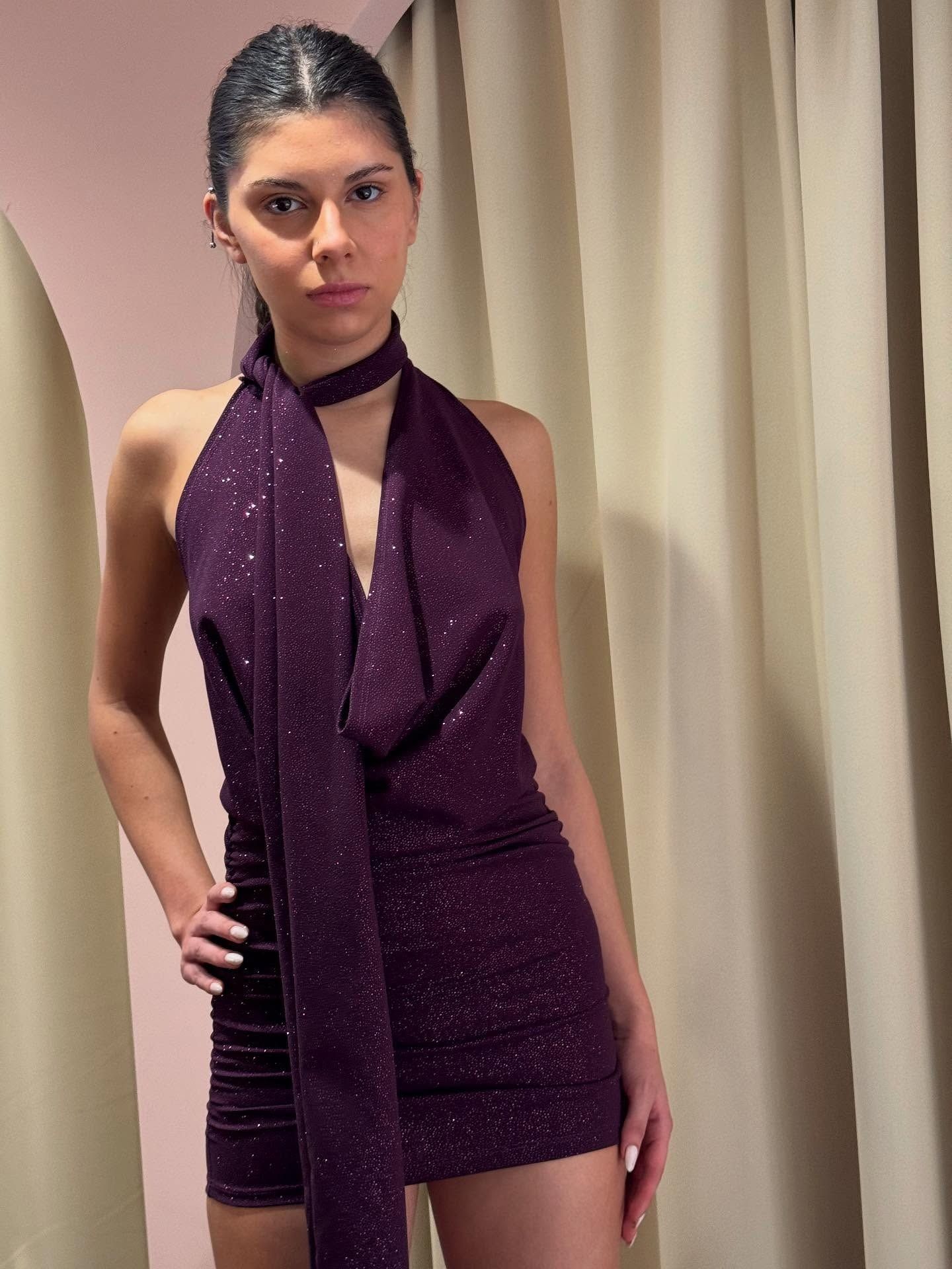 Mujer con un vestido morado brillante y bufanda, de pie con la mano en la cadera en una habitación con tela drapeada.