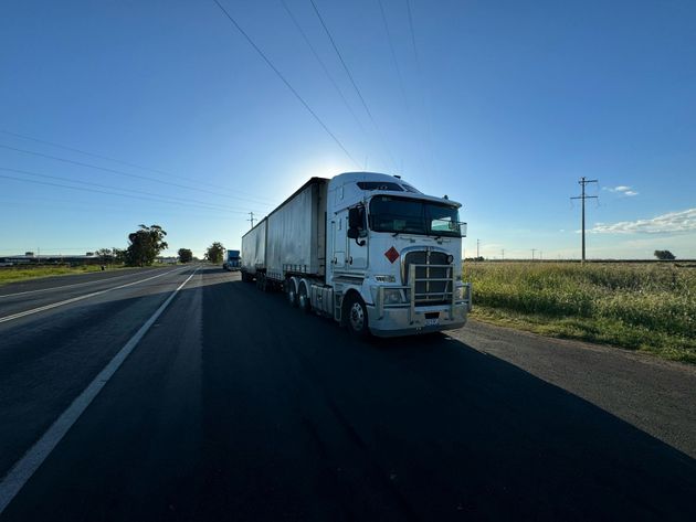 White semi-truck driving on a rural highway beside open fields under a clear blue sky