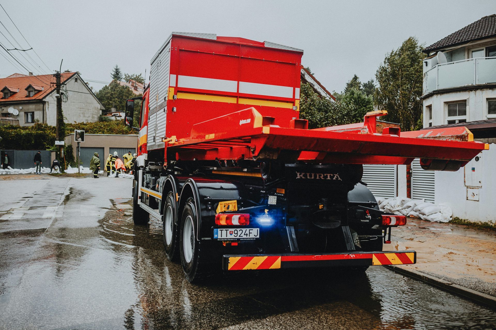 Red tow truck on a wet residential street, hauling a red trailer past houses after rain.