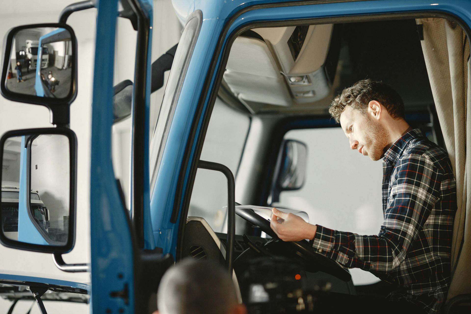 Man in plaid shirt working inside a blue truck cab, looking at a device near the dashboard.