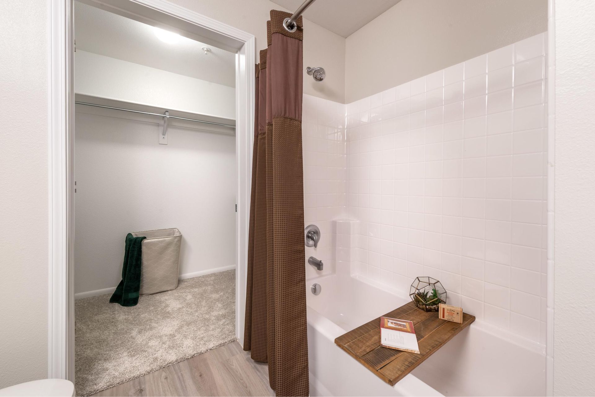 Bathroom with white tile tub/shower, brown curtain, and a wood shelf; doorway to a carpeted closet with hanging rod.