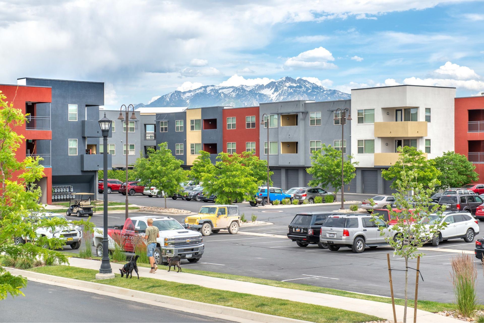 People walking two dogs near a colorful apartment complex exterior with a parking lot.