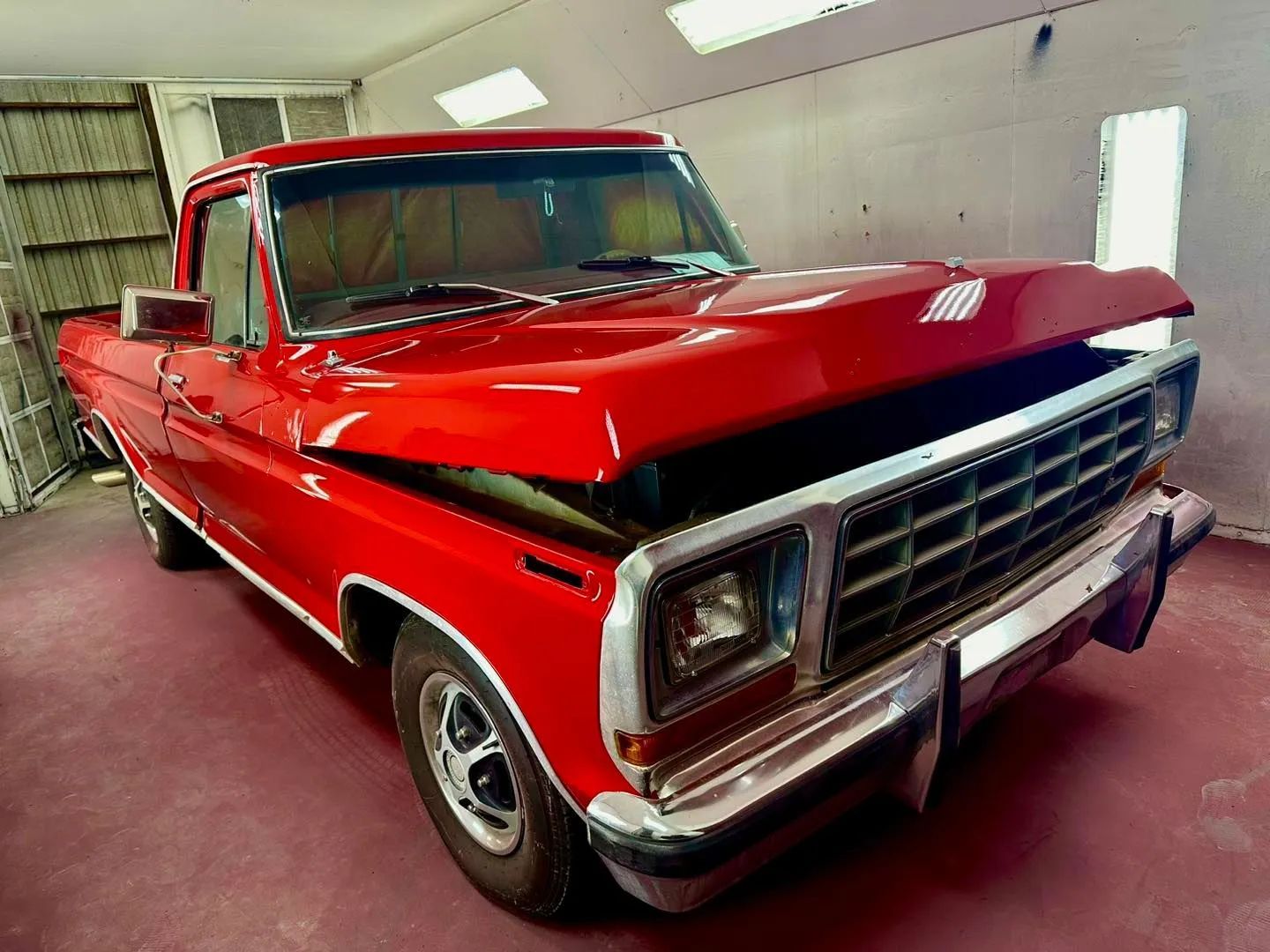 Red classic pickup truck in a paint booth with open hood.