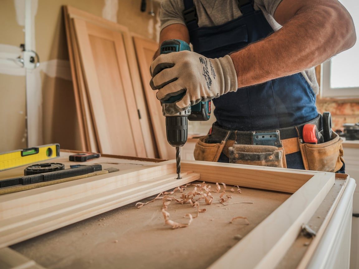 Carpenter drilling wood with a power drill; sawdust present.