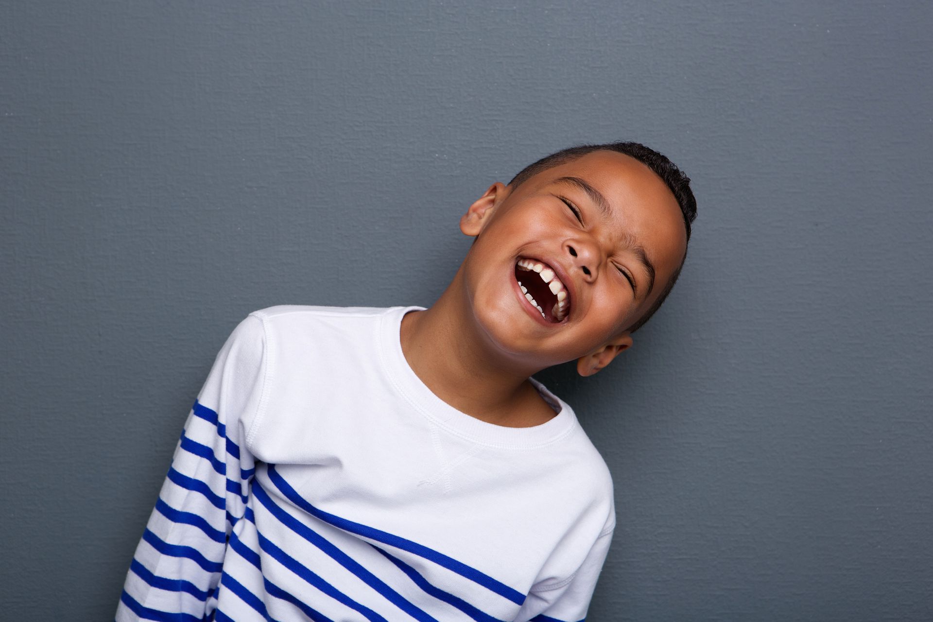 Boy laughing with his head tilted, wearing a blue and white striped shirt. Against a gray background.