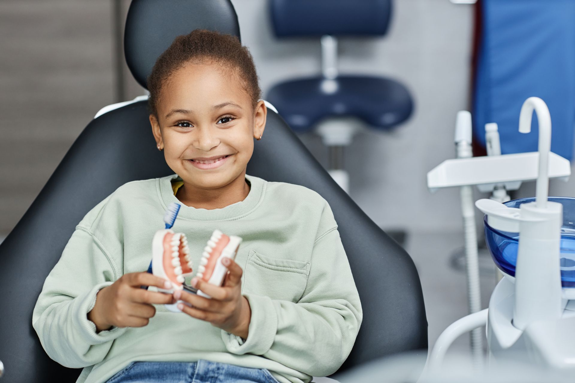 Girl in a dental chair holding a model of teeth, smiling.