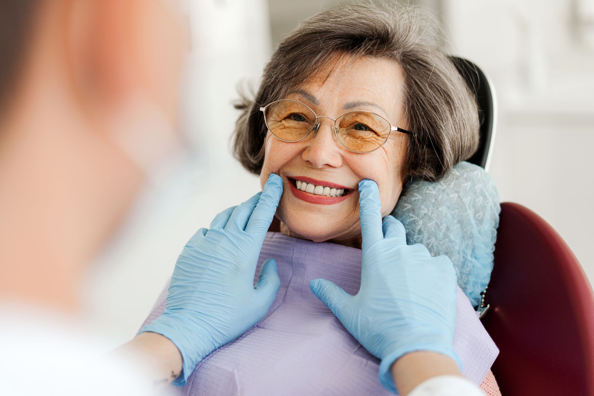 Dentist examining patient's smile in dental chair; dentist's gloved hands gently touch patient's cheeks.
