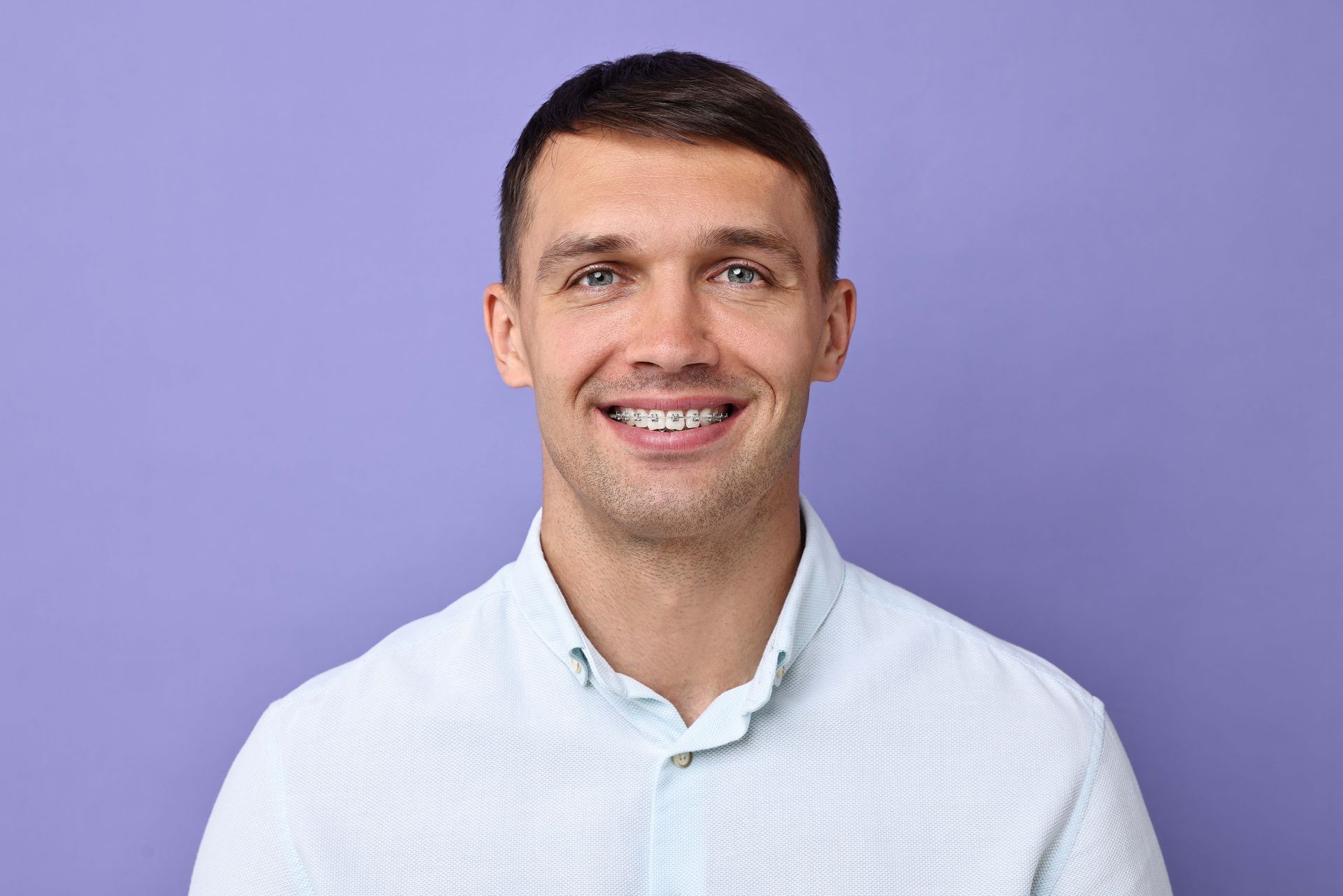 Man with short hair and braces smiles, wearing a light blue button-down shirt, against a purple background.