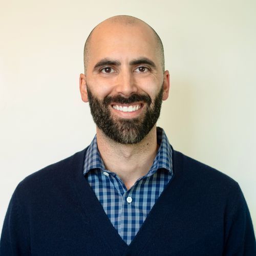 A bald man with a beard is wearing a blue sweater and a plaid shirt.