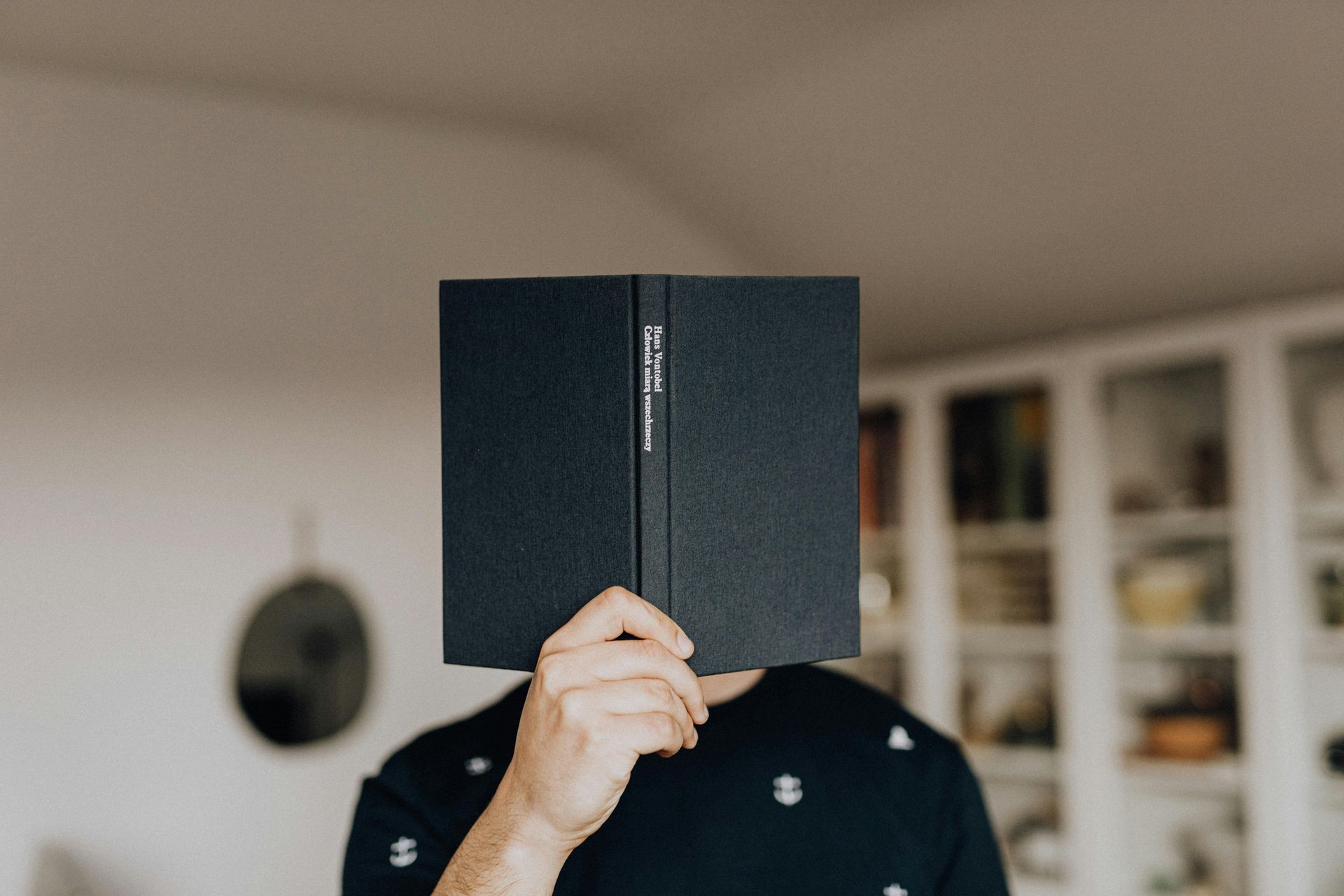 A person holds a dark book in front of them, the book obscuring their head entirely.