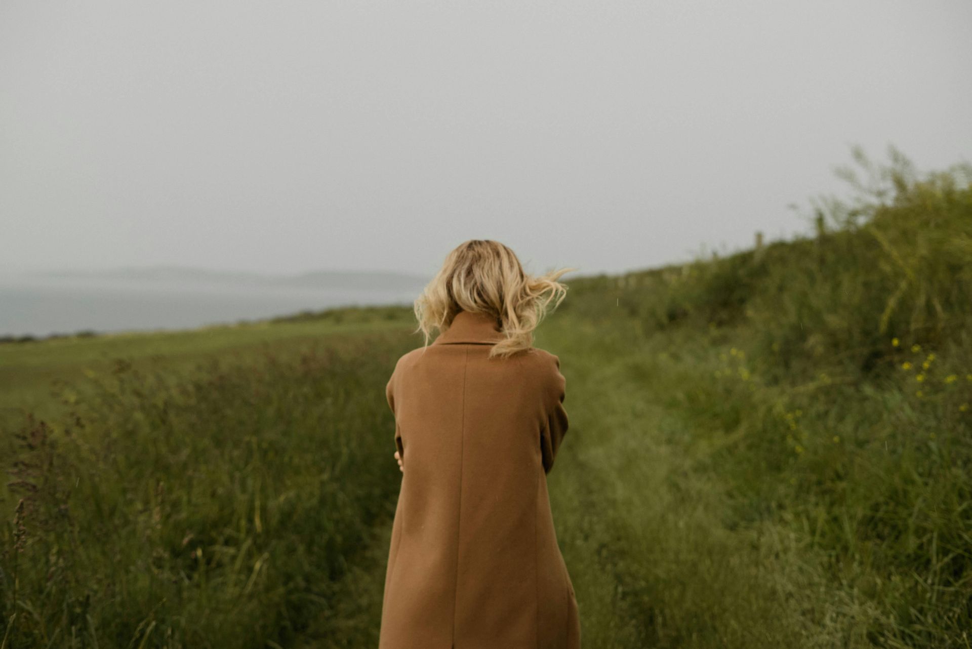 Woman in brown coat stands in field, facing away, overlooking water.