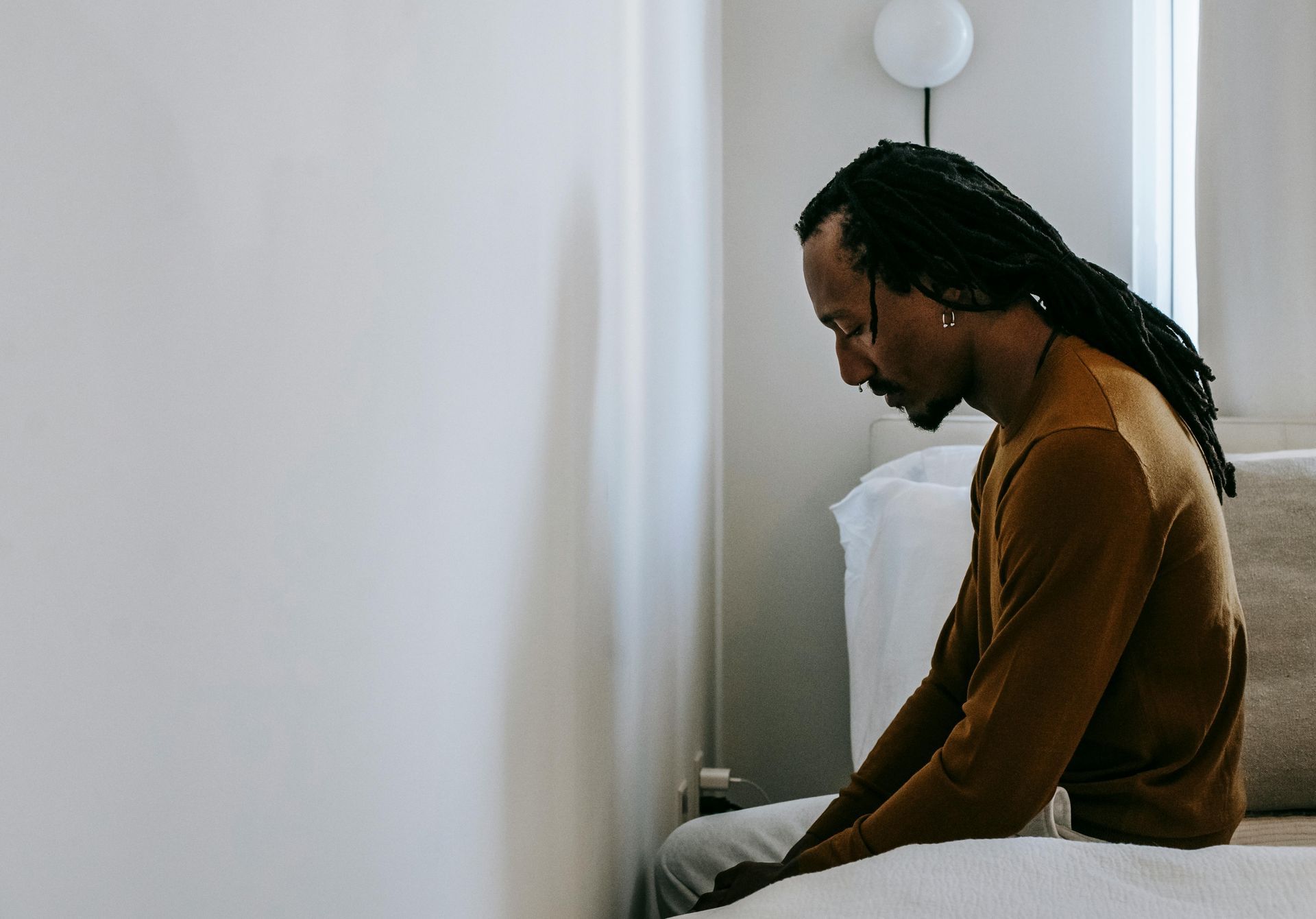 A person sits at the edge of a bed, slumped forward, seeming despondent.  A person sits at the edge of a bed, slumped forward, seeming despondent.