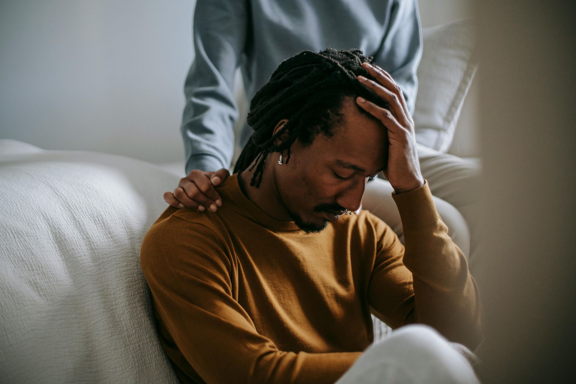 A grieving person sits with head bowed and in hand. Behind them, another person rests a comforting hand on their shoulder.  A grieving person sits with head bowed and in hand. Behind them, another person rests a comforting hand on their shoulder.