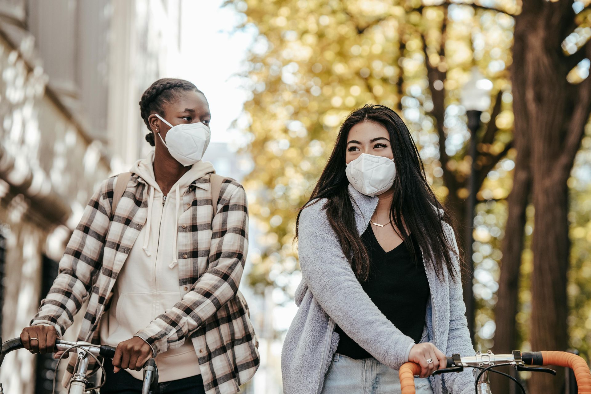 Two people wearing KN95 respirators walk their bicycles together down a sunny autumn street, appearing happy.  Two people wearing KN95 respirators walk their bicycles together down a sunny autumn street, appearing happy.