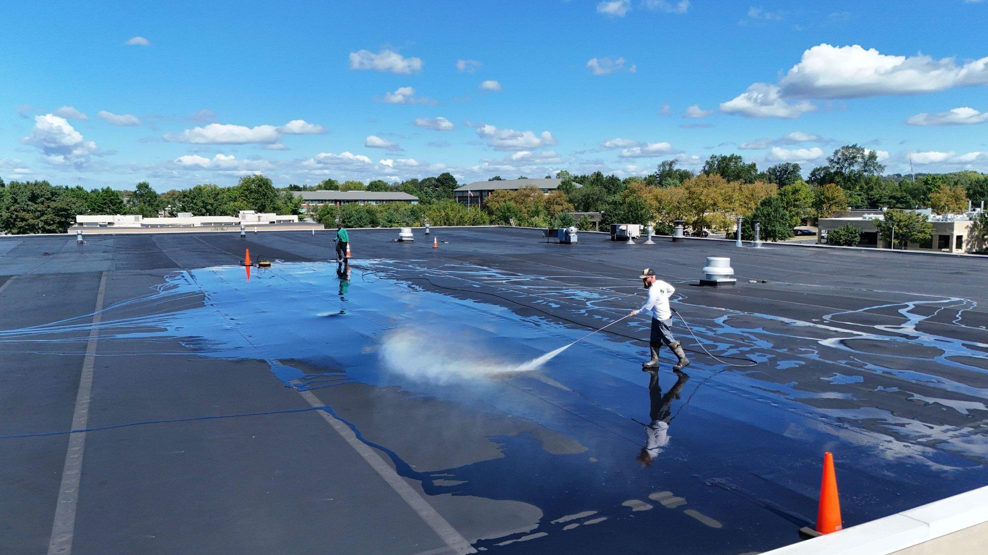 Two people on a flat roof, spraying water. Large puddles reflect the blue sky. Orange cones mark the perimeter.