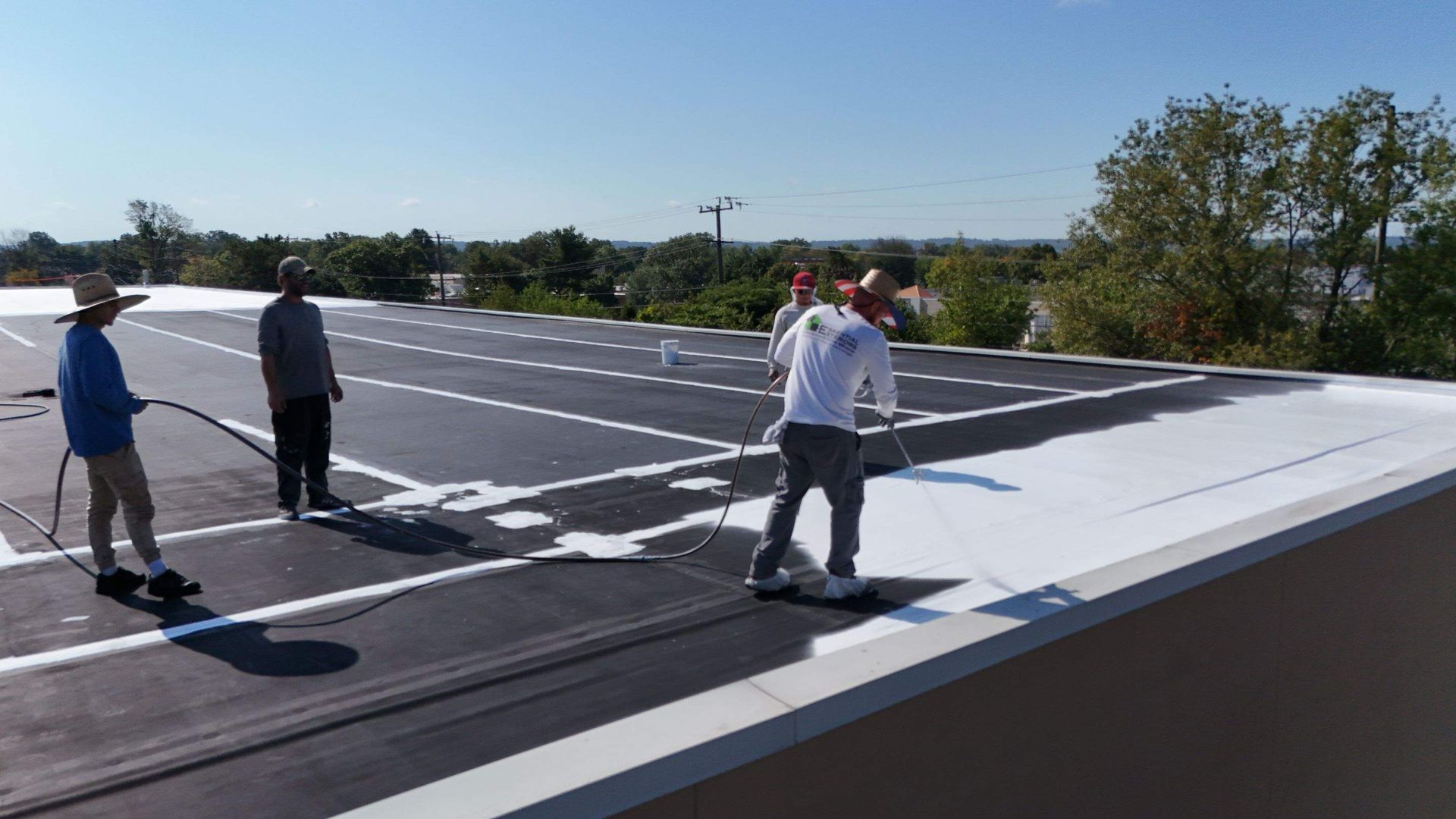 Workers applying white coating to a flat roof with sprayers on a sunny day.