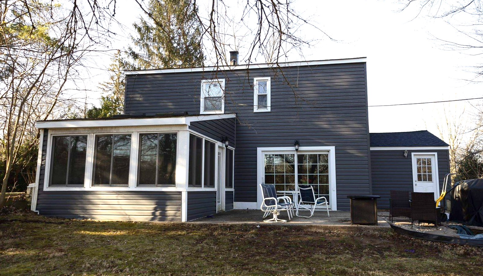 A two-story dark gray house with a white-framed sunroom, sliding glass doors, and a backyard patio under a clear sky.