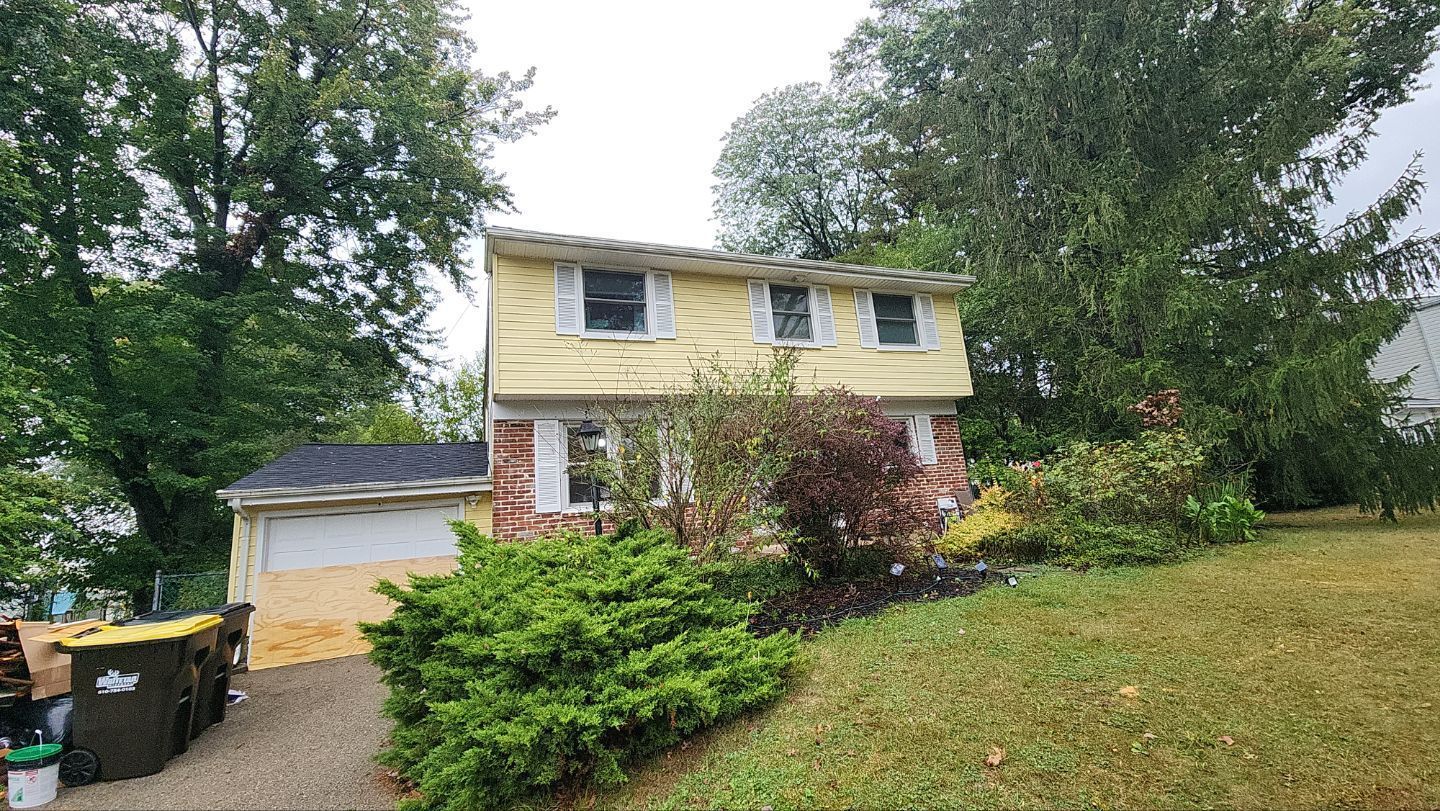 A two-story yellow house with brick accents, partially obscured by trees and a bush, beside a gravel driveway and bins.
