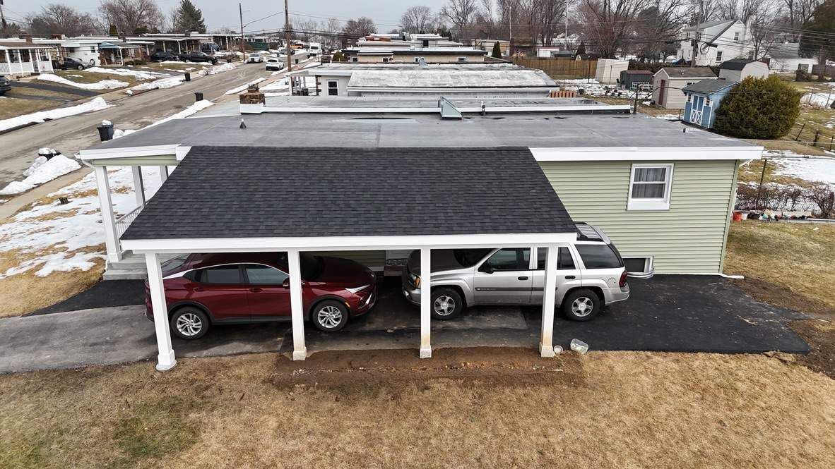 An aerial view of a pale green single-story home with a black-roofed carport containing a maroon and a silver SUV.