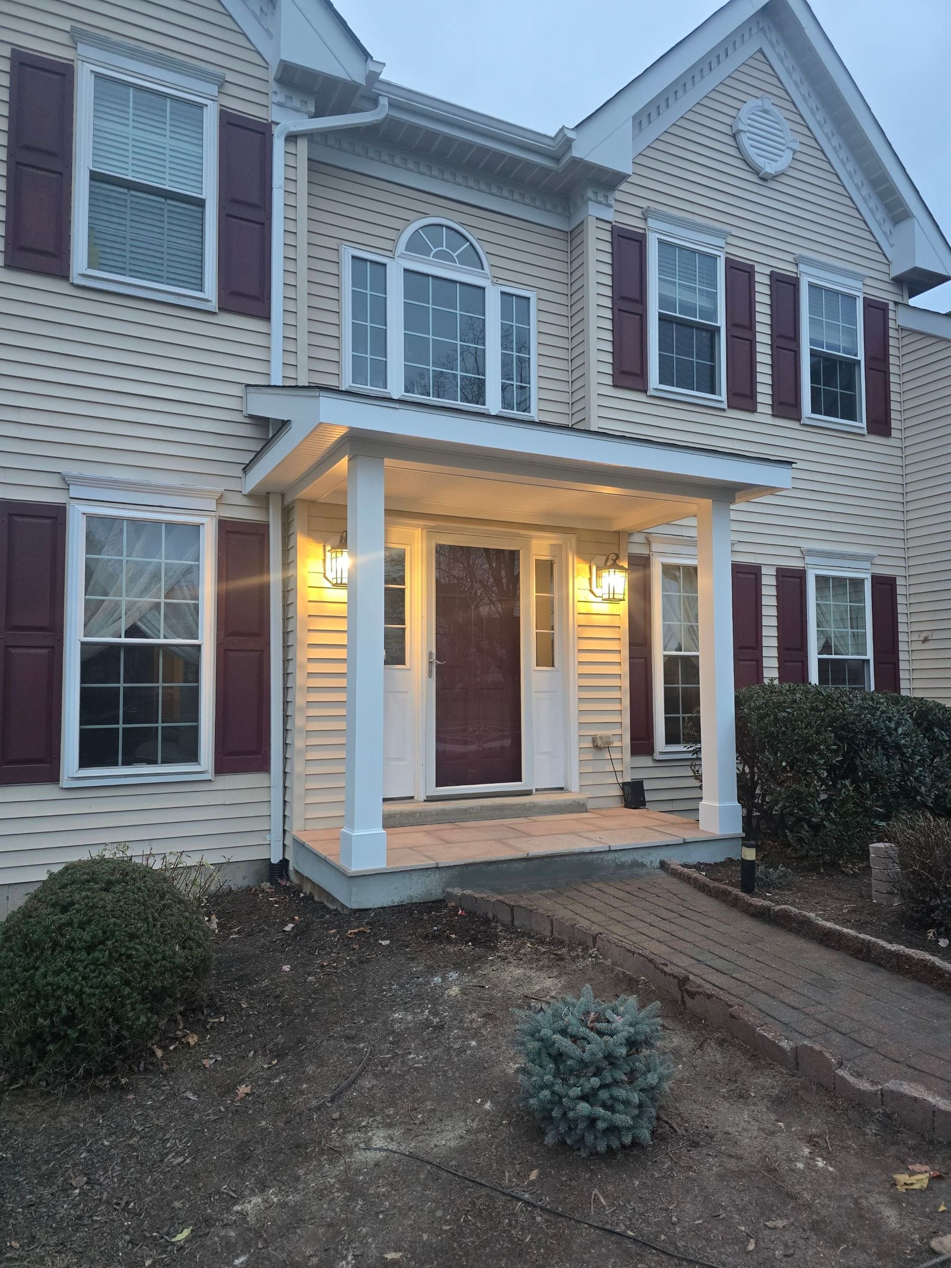 A two-story, light yellow house with a covered front porch, dark red shutters, and a brick walkway leading to the entry.