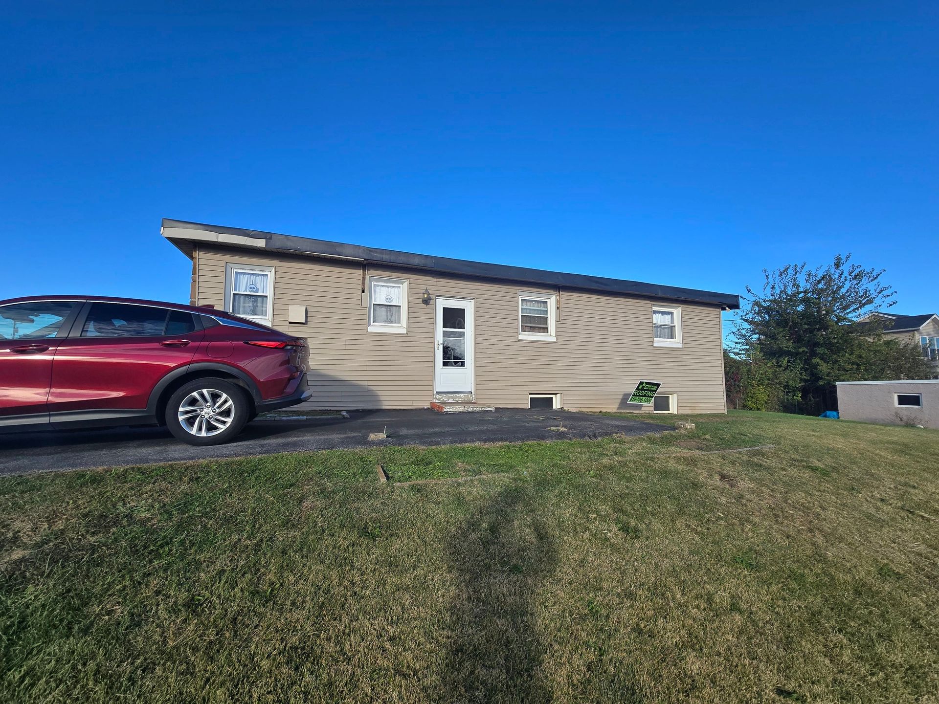 A single-story, tan-sided house with a shed roof, a central white door, and a red car parked on an asphalt driveway.