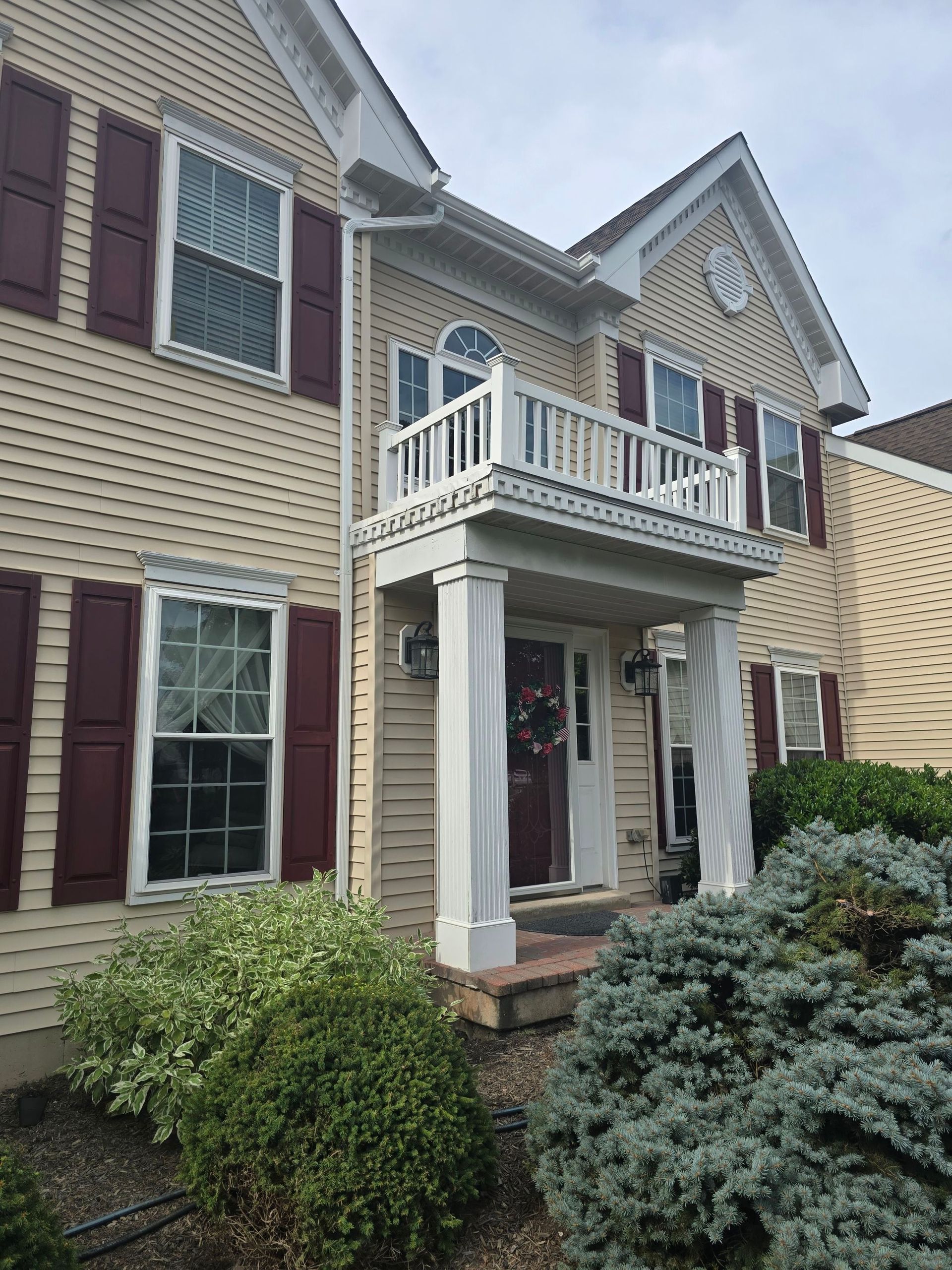A two-story tan suburban house featuring a front porch with a small second-floor balcony, white pillars, and maroon shutters.