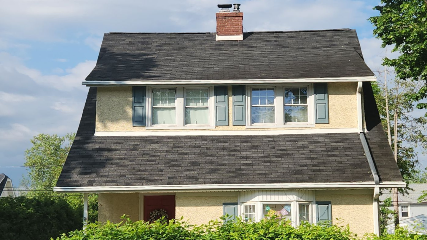 A two-story tan house with a gambrel roof, a brick chimney, and dark green shutters on a sunny day.