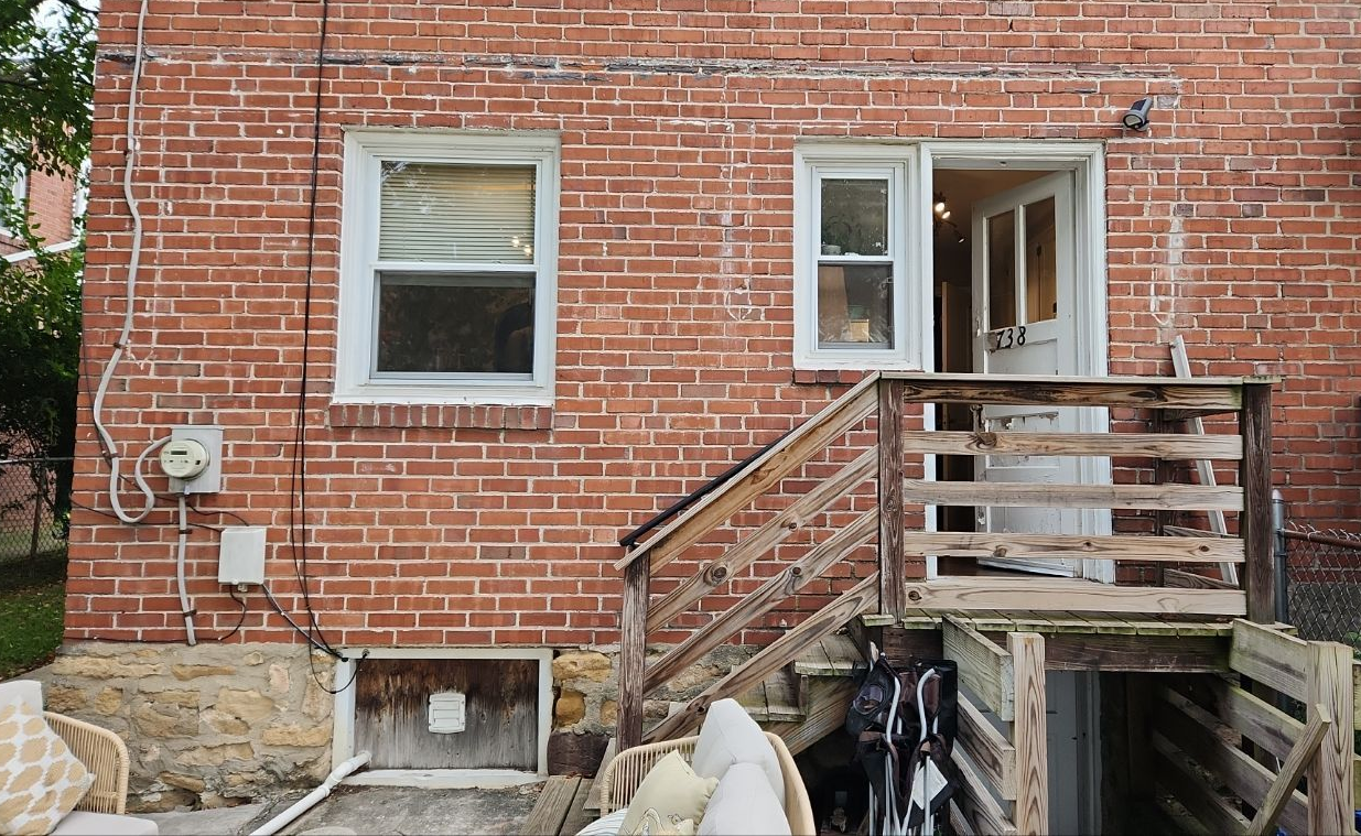 Back exterior of a brick house featuring a small wooden porch, stairs leading to a doorway, and a basement window.