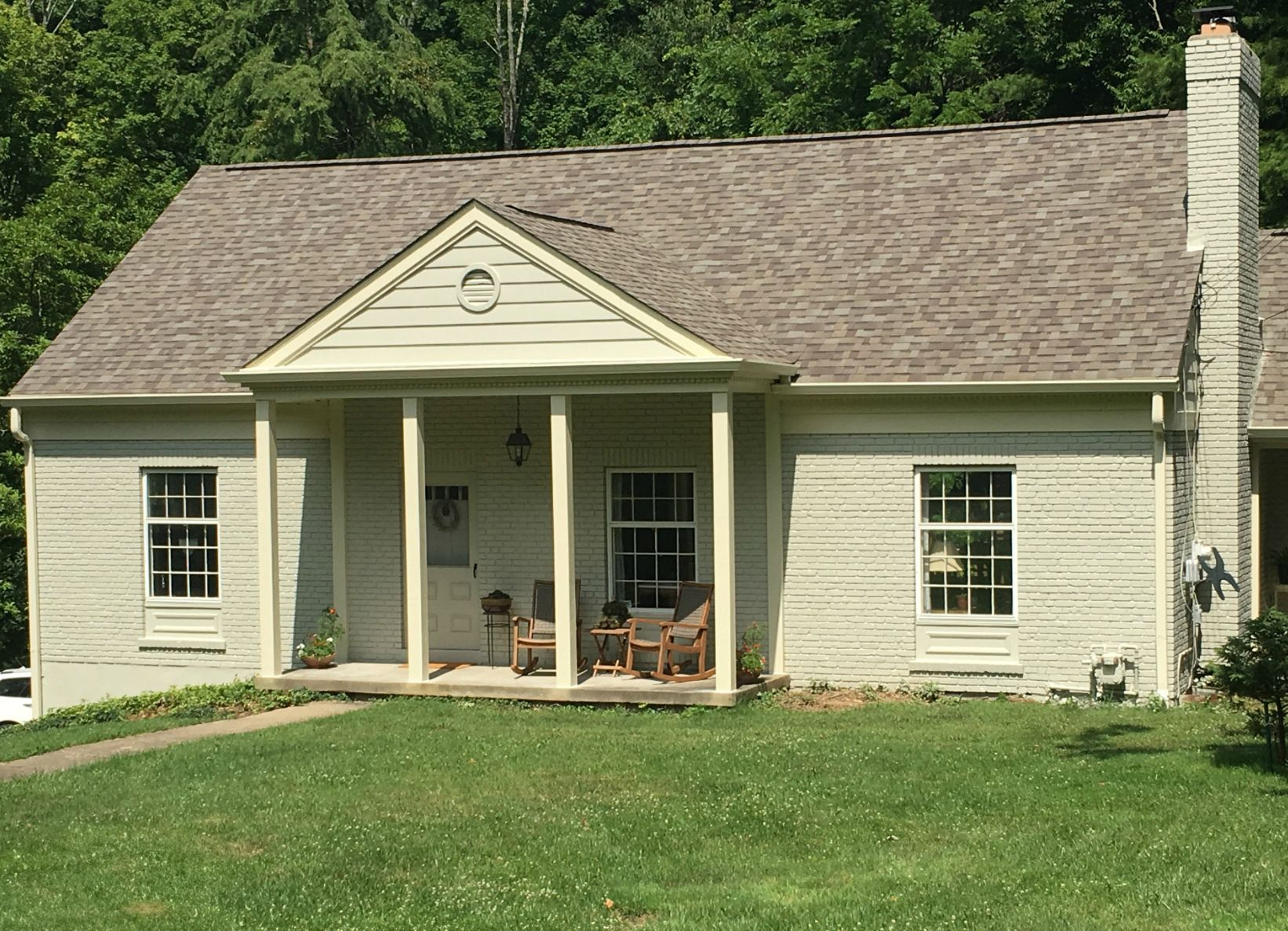 A single-story, light-colored house with a covered front porch, stone chimney, and a brown shingled roof.