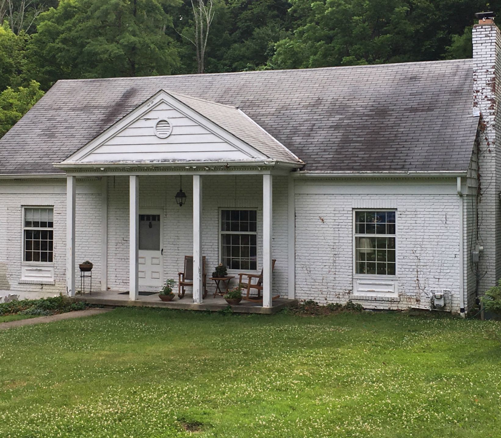 A small, white, single-story house with a gabled porch, green lawn, and dark roof, surrounded by trees.