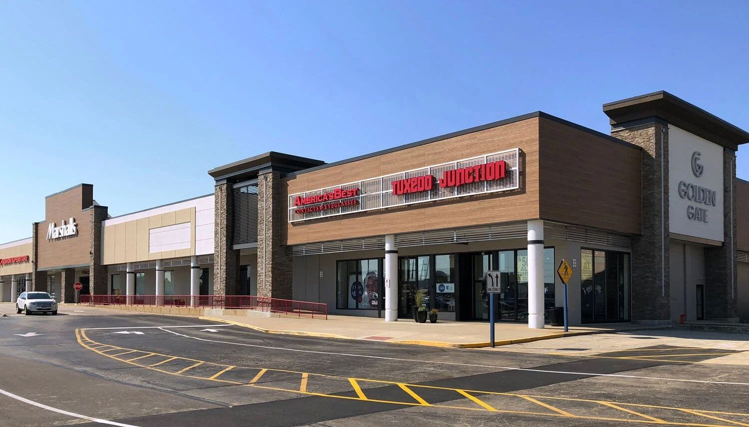 A strip mall with various store fronts under a clear blue sky. Stores include a bakery, a fitness center, and a cafe.