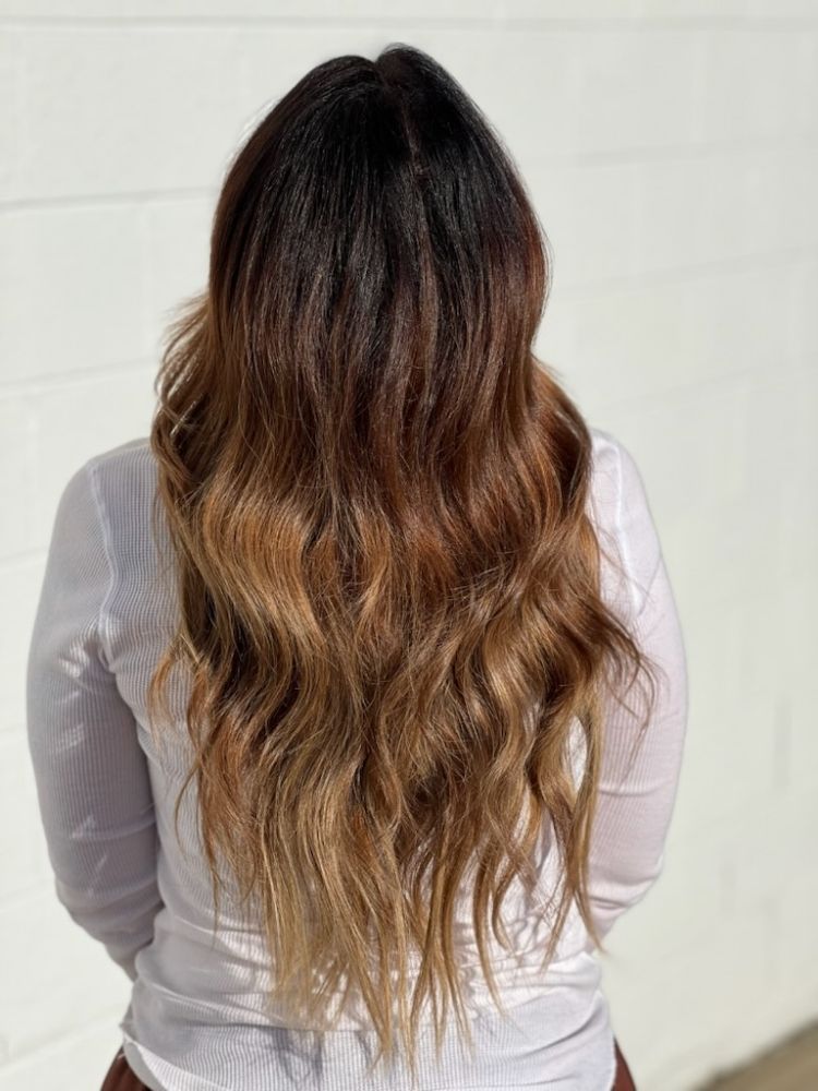 A woman with long wavy brown hair is standing in front of a white wall.