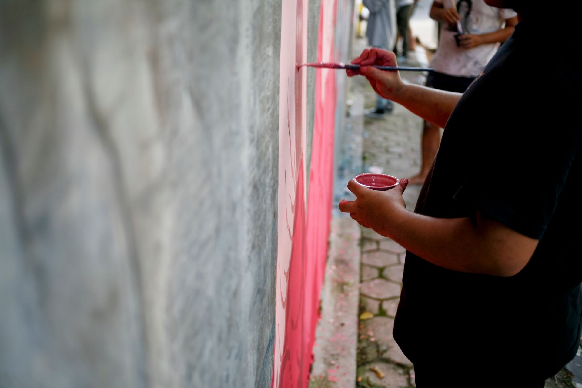 Person painting a pink stripe on a gray wall with a paintbrush and holding a paint container outdoors.