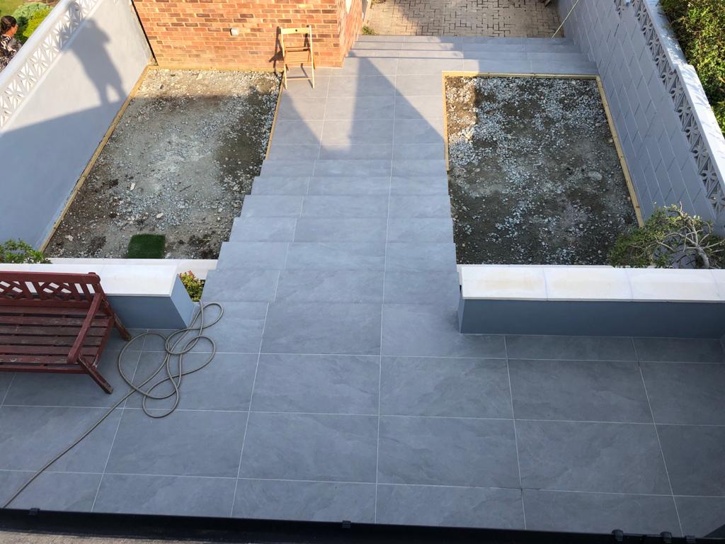 A wooden bench is sitting on a tiled patio next to a brick wall.