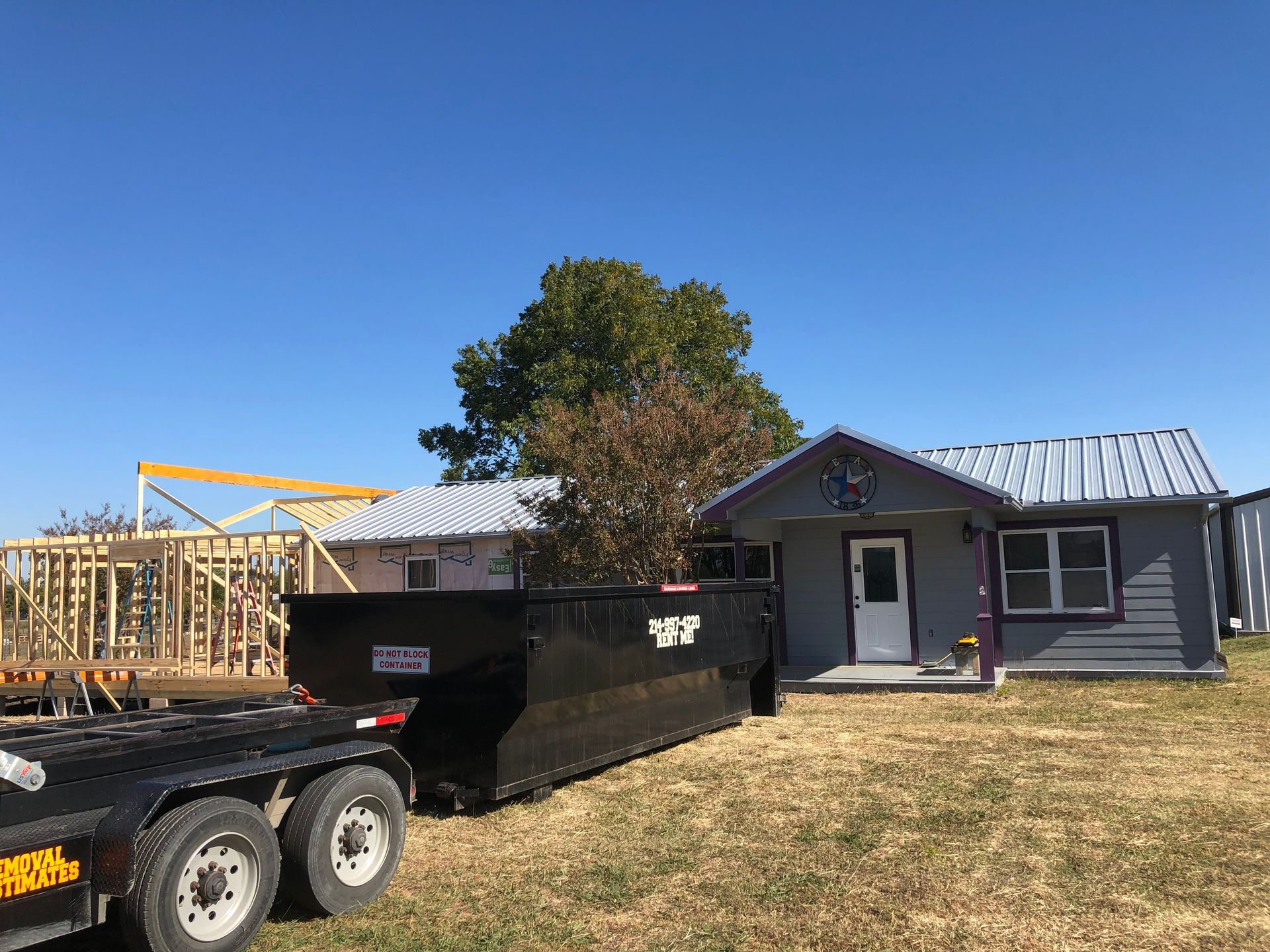 Black Iron Manroll-off dumpster at a construction site with a house and partial wood frame structure